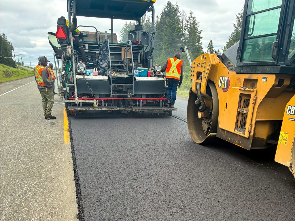 Dawson Construction paving crew laying fresh asphalt on Highway 97 near Lac La Hache, with a CAT roller compacting behind the paver