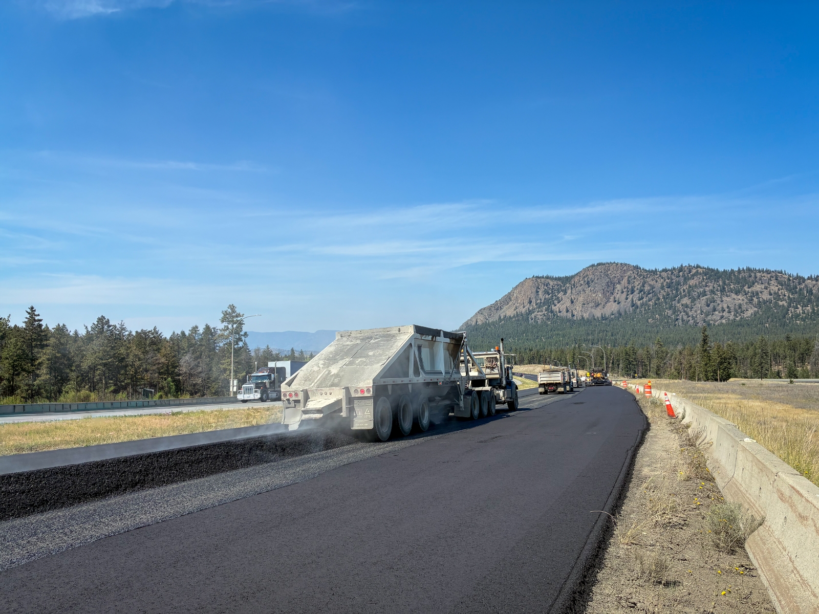 Paving train of trucks and equipment laying asphalt on Highway 5 near Kamloops with mountains in the background